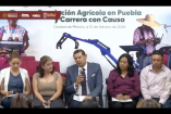 Alejandro Armenta habla durante la presentación de la Revolución Agrícola en Puebla y la Carrera con Causa, acompañado por funcionarios, con imágenes alusivas al campo y maquinaria agrícola de fondo.