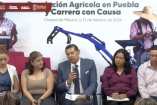 Alejandro Armenta habla durante la presentación de la Revolución Agrícola en Puebla y la Carrera con Causa, acompañado por funcionarios, con imágenes alusivas al campo y maquinaria agrícola de fondo.