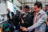 Estudiante explica proyecto tecnológico con laptop a autoridad educativa durante actividad de educación dual en espacio de exhibición con docentes.