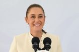 Claudia Sheinbaum sonriendo durante su conferencia mañanera en Tabasco, de pie detrás de un atril presidencial con el Escudo Nacional y la bandera de México de fondo.
