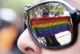 Una persona con una bandera LGBTTTIQ+ se refleja en unos anteojos obscuros.