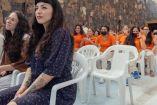 Mon Laferte con vestido azul sentada en silla de plástico frente a mujeres de penal