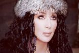 Cher con sombrero peludo café y cabello rizado, usa vestido negro