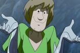 Shaggy con playera verde con manos arriba y sonriendo, rocas de fondo