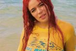 Karol G con camiseta amarilla mojada; sobre la arena y mar atrás