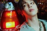 Jimin con cabello en frente, collar de caracoles recargado en cajuela de automóvil