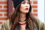 Megan Fox con gorro negro y chamarra verde; pared de ladrillos atrás