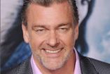 Ray Stevenson sonriendo con traje gris y camisa rosa