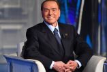 Silvio Berlusconi sentado en sillón presidencial sonría con sorna 