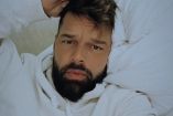 Ricky Martin con sudadera blanca y mano en cabeza, acostado en cama