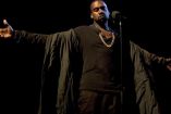 Kanye West con playera y abrigo café, extendiendo brazos enfrente de micrófoni