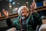 Ida Vitale, poeta uruguaya de cabello blanco con gafas, sentada en un espacio con libros e imágenes, haciendo gestos expresivos con sus manos en su 100º cumpleaños