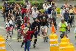 La imagen muestra un grupo diverso de personas montando bicicletas en un evento al aire libre. Se observan ciclistas de diferentes edades y atuendos, desde los que llevan equipo de seguridad completo, como cascos y rodilleras, hasta aquellos en ropa casual. Una mujer con una sudadera roja y una gorra está sosteniendo una señal de alto, guiando a los ciclistas por el recorrido. La imagen captura un momento de actividad comunitaria, promoviendo el ciclismo como una forma saludable y ecológica de transporte y 