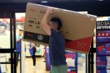 Una persona lleva una caja grande de una televisión 4K UHD sobre su hombro, navegando por un centro comercial con señalizaciones de descuentos de El Buen Fin