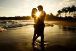 Una pareja se abraza amorosamente en una playa al atardecer, con el sol creando un brillante contorno a su alrededor. El cielo está teñido de naranja y amarillo, y hay palmeras en el fondo, creando un escenario romántico y sereno.