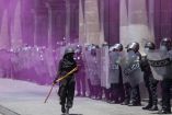 Manifestante encapuchado, armado con un palo, enfrenta una fila de policías antidisturbios entre una nube de humo morado, en un poderoso momento de tensión civil.