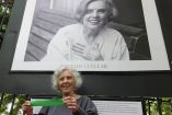 Elena Poniatowska, de pie y sonriente, sostiene una cinta verde mientras se encuentra frente a una gran fotografía de ella misma en la Galería Abierta de Chapultepec, capturada por Rogelio Cuéllar, como parte de un homenaje a su legado literario y cultural.