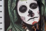 Mural de una Catrina en una pared de la ciudad, decorada con elementos tradicionales del Día de Muertos, mientras dos personas caminan al frente del mural.