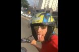 Motociclista joven tomando una selfie con casco y gafas de sol mientras conduce por una vía urbana en São Paulo, Brasil.