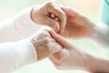 Manos de una persona adulta mayor tomadas con delicadeza por una persona más joven en un entorno cálido y luminoso, simbolizando apoyo emocional y cuidado en el contexto del Parkinson y enfermedades neurodegenerativas