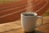 Taza de café humeante sobre una mesa de madera junto a una pista de atletismo, simbolizando la relación entre el consumo de café y el aumento del rendimiento físico y mental en el deporte.