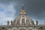 Cúpula de la Basílica de San Pedro en el Vaticano con cielo nublado y estatuas de santos en primer plano.
