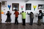 Grupo de adultos mayores realiza ejercicios de rehabilitación apoyándose en una pared dentro de un centro comunitario. Actividades dirigidas a mejorar el equilibrio y la movilidad, comunes en terapias para personas con párkinson u otras afecciones neurológicas. Decoraciones y arte en el fondo.