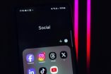 Primer plano de un teléfono móvil con una carpeta titulada "Social", que muestra íconos de Facebook, Instagram, Threads, X, LinkedIn, TikTok y YouTube, con fondo oscuro y luces de neón rosas. La imagen ilustra visualmente la presencia de diversas redes sociales en dispositivos móviles en España en 2025