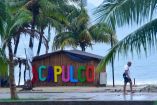 Letrero multicolor de “ACAPULCO” en un quiosco de caña junto al mar, palmeras húmedas y un hombre caminado descalzo sobre el malecón mojado por la tormenta Flossie; el cielo nublado y la arena reflejan el agua de la lluvia.