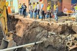 Un grupo de funcionarios y trabajadores con chalecos de seguridad inspeccionan un socavón de gran profundidad que se abrió en la colonia La Duraznera, Tlaquepaque, Jalisco. En la imagen se observa maquinaria pesada, tuberías rotas y daños estructurales en la banqueta, evidenciando la magnitud del hundimiento.