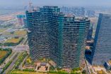 Vista aérea del Regent International, un rascacielos de forma ondulada ubicado en Hangzhou, China, reconocido por ser el edificio residencial con mayor número de habitantes en el mundo. La imagen muestra su estructura de vidrio y la alta densidad de balcones ocupados, rodeado de otras construcciones modernas y zonas verdes urbanas.