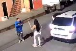 Captura de una cámara de seguridad que muestra el momento previo al feminicidio de Karla Bañuelos en una calle de Guadalajara. En la imagen se observa a una mujer enfrentando a un hombre con sudadera azul que porta un arma larga, frente a una camioneta blanca. La escena ocurre a plena luz del día.