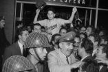 Ángel Macías, vestido con el uniforme de la Liga Industrial de Monterrey, es cargado en hombros y ovacionado por una multitud al llegar a la ciudad tras lanzar un juego perfecto en la final de la Little League en 1957. La imagen muestra la euforia colectiva, con soldados y civiles celebrando su regreso en una estación marcada con el letrero "Sala de Espera"
