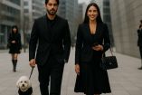 Pareja joven caminando por una ciudad moderna, ambos vestidos completamente de negro. Él lleva un suéter negro y pasea un perro negro, mientras ella viste un conjunto formal con falda y saco, sonriendo con un celular en la mano. La imagen transmite elegancia, sobriedad y un estilo urbano contemporáneo, bajo un cielo nublado.