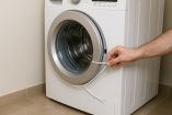 Mano masculina usando un cordón blanco para intentar desbloquear la puerta de una lavadora de carga frontal. El cordón rodea el aro de la puerta mientras se aplica presión hacia el cierre, en un entorno doméstico con piso de cerámica.
