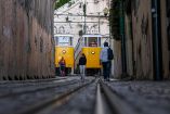 Dos funiculares amarillos del sistema Lavra, detenidos y fuera de servicio tras el accidente del funicular de la Gloria en Lisboa. En primer plano, personas caminan por las vías mojadas en una calle estrecha entre edificios. La imagen refleja el impacto del cierre del servicio turístico tras la tragedia.