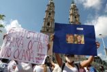 Manifestantes en Tabasco sostienen carteles con mensajes de justicia y fotos de Octavio Ocaña durante una protesta pacífica frente a una iglesia de arquitectura colonial; algunos portan cubrebocas, con cielo parcialmente nublado