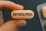 Píldora de color salmón con la palabra "Orforglipron" impresa, sostenida entre los dedos de una mano, con un fondo desenfocado que incluye un blíster de tabletas. Representación conceptual del medicamento oral desarrollado por Eli Lilly para el tratamiento de la obesidad.