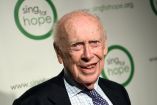 James Watson, co-descubridor de la estructura del ADN y ganador del Premio Nobel de Medicina, posa durante un evento de la organización Sing for Hope en Nueva York, vistiendo traje oscuro y corbata, con fondo institucional del evento.
