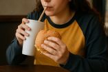 Mujer embarazada sosteniendo una hamburguesa y un vaso con bebida, representando los antojos por comida ultraprocesada durante el embarazo, asociados a factores emocionales y posibles riesgos para la salud materna y fetal.