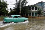 Un automóvil clásico color verde claro circula por una calle completamente inundada en una zona urbana de La Habana, Cuba. Al fondo se observan edificios antiguos y palmas, reflejando la situación de infraestructura deteriorada y las frecuentes inundaciones que afectan a la capital cubana.