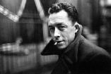 Retrato en blanco y negro de Albert Camus mirando a cámara, con abrigo oscuro y un cigarrillo en la boca, en un entorno urbano desenfocado que refuerza su imagen de escritor reflexivo y existencialista.