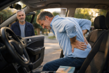 Hombre de mediana edad con gesto de dolor lumbar al salir de un coche, mientras otra persona lo observa, una escena cotidiana que ilustra las primeras señales de aviso del cuerpo antes de que el dolor de espalda se vuelva crónico.