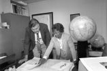 Gladys West, matemática estadounidense y pionera del GPS, revisa mapas y documentos técnicos junto a un colega en una oficina, con globos terráqueos y material cartográfico al fondo.