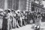 Fotografía histórica en blanco y negro de un grupo numeroso de trabajadores rurales formados en fila frente a un edificio de piedra, mientras una mujer organiza o reparte atención, escena representativa del contexto social y laboral en México.