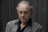 Retrato de Frederick Wiseman, documentalista estadounidense, con saco gris y camisa oscura, frente a un fondo textil. La imagen acompaña una nota sobre su fallecimiento a los 96 años y su legado en el cine documental.