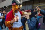 Joven con gorro de cabeza de perro y chamarra roja interactúa con joven que porta máscara de dinosaurio durante la reunión therian en la Explanada de Rectoría de la UANL, ante la mirada de otros estudiantes.