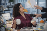 Mujer en oficina con un croissant en la boca mientras sostiene café y un biberón, recibe documentos y atiende tareas laborales frente a una computadora, imagen que simboliza la carga mental y la doble jornada de las madres trabajadoras.