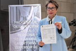Delia González Cobos sostiene el documento de registro como aspirante a encabezar la Auditoría Superior de la Federación, frente a un cartel del proceso de designación en la Cámara de Diputados.