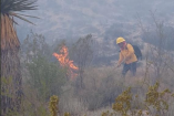 Elemento de brigada forestal con casco amarillo y equipo de protección trabaja para sofocar llamas activas en zona de matorral y pastizal en Múzquiz, Coahuila, en medio de humo denso por incendio forestal.